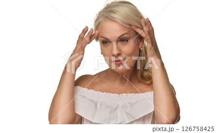 Senior woman gently touches temples while evaluating skin with focused expression against white background, showing beauty and self-acceptance Senior woman gently touches temples while evaluating skin with focused expression against white background, showing beauty and self-acceptance 126758425