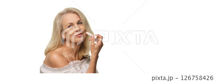 Elegant senior woman holding lip balm and smiling confidently with soft expression against white background in off-shoulder blouse 126758426