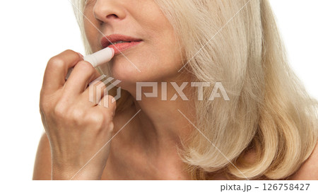 Elderly woman softly applies lip balm to lips with gentle motion, showing hydrated skin and soft glow against white studio background Elderly woman softly applies lip balm to lips with gentle motion, showing hydrated skin and soft glow against white studio background 126758427