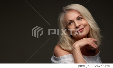 Senior woman smiling gently and resting chin on hand against dark background showing youthful glow and soft lines Senior woman smiling gently and resting chin on hand against dark background showing youthful glow and soft lines 126758460