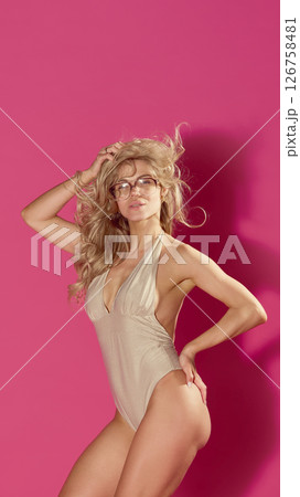 Energetic portrait of beautiful smiling blonde woman posing in beige retro stylish swimsuit and holding hand on hip, hair flowing on wind against pink background Energetic portrait of beautiful smiling blonde woman posing in beige retro stylish swimsuit and holding hand on hip, hair flowing on wind against pink background 126758481