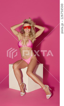 Smiling woman in pink bikini lifts hands behind head while sitting on cube in red sunglasses against pink background Smiling woman in pink bikini lifts hands behind head while sitting on cube in red sunglasses against pink background 126758500