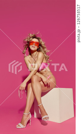 Blonde woman in pink bikini and red sunglasses leans forward sitting on cube with crossed legs and confident expression against pink background Blonde woman in pink bikini and red sunglasses leans forward sitting on cube with crossed legs and confident expression against pink background 126758517