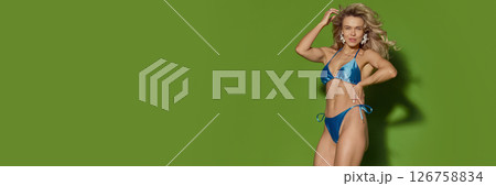 Smiling blonde woman, wearing in blue bikini posing with hand on hip against green background. Banner with copy space 126758834
