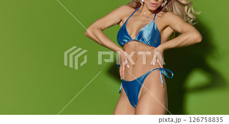 Close-up portrait of sensual blonde woman in shiny blue bikini with hands on waist and wavy hair against green studio background. 126758835