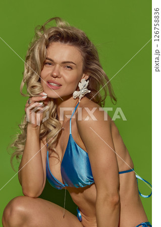 Close-up portrait of sensual blonde woman in shiny blue bikini looking at camera with confidence against green studio background. 126758836
