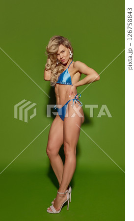 Blonde woman in shiny blue bikini and white heels standing confidently with hand on waist against green background 126758843