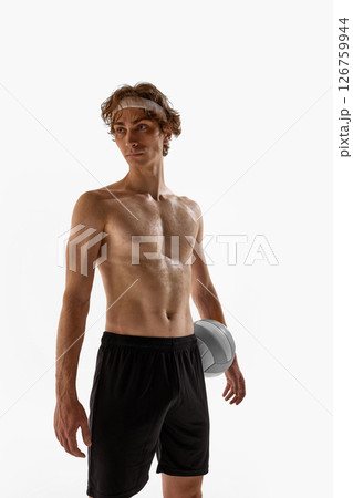 Shirtless male athlete with confident look holds volleyball at waist while standing tall in black shorts against white studio background. 126759944