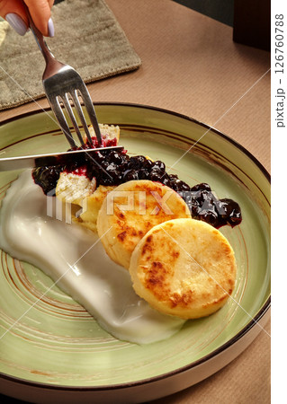 Woman eating syrniki with blueberry jam and sour cream using fork and knife 126760788