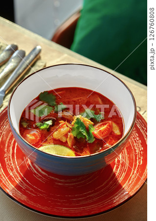 Spicy thai tom yum soup is served in blue bowl on red plate in restaurant Spicy thai tom yum soup is served in blue bowl on red plate in restaurant 126760808