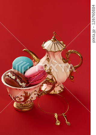 Colorful macarons resting in elegant teacup beside decorative teapot on red background 126760884