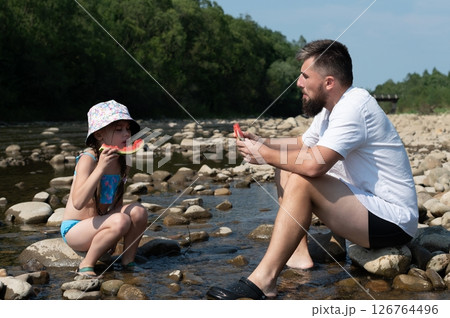 Father and daughter enjoy a refreshing watermelon snack by the river on a sunny day. Father and daughter enjoy a refreshing watermelon snack by the river on a sunny day. 126764496