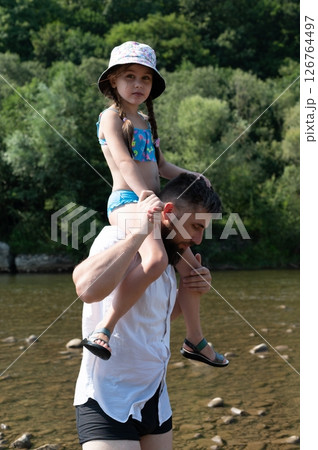 A father carries his daughter on his shoulders by a river. A father carries his daughter on his shoulders by a river. 126764497