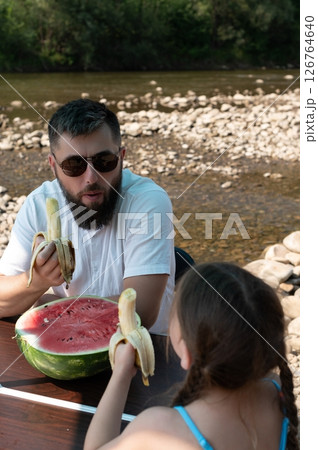 A father and daughter enjoy a picnic by the river, sharing watermelon and bananas. 126764640