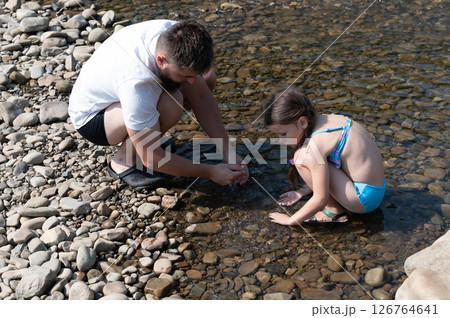 A father and daughter enjoy a sunny day playing in a shallow stream, splashing water and exploring the smooth river stones. A father and daughter enjoy a sunny day playing in a shallow stream, splashing water and exploring the smooth river stones. 126764641