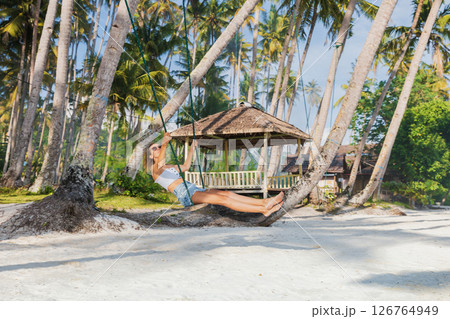 Woman on swings at tropical beach, enjoying freedom and sunshine under palm trees, paradise vibes. 126764949