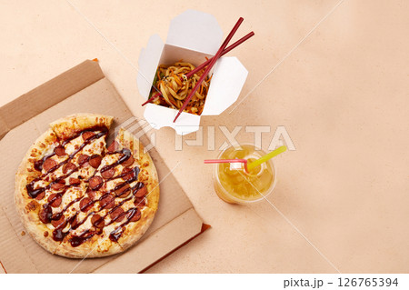 A tempting round pizza topped with cheese and slices of pepperoni sits next to a takeout container filled with colorful noodles. 126765394