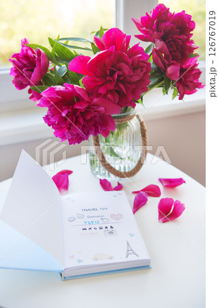 A travel journal open to a page with stickers about a trip to Paris sits on a white table next to a glass vase filled with peony flowers, hinting at romantic travel 126767019