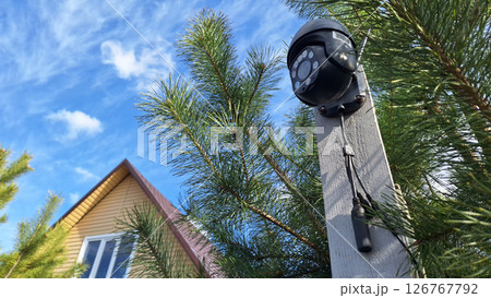 Outdoor security camera on wooden post near house and pine trees under blue sky 126767792