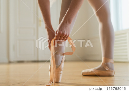 Ballerina puts on pointe shoes on leg in dance class. Classical ballet dancer woman in dance studio. Ballerina putting on tying classical pointe shoes for dance training. Ballet school concept 126768046