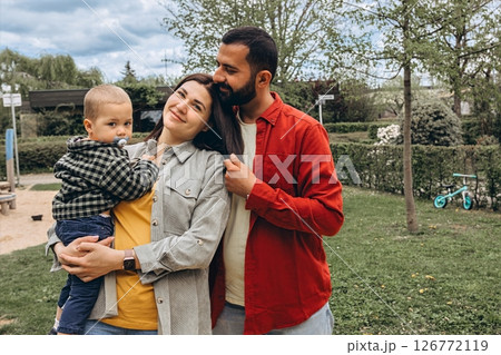 Young family spending time together outdoors in a park. Mother holds a toddler while father embraces them, all dressed in casual clothes, enjoying a peaceful day in nature. Young family spending time together outdoors in a park. Mother holds a toddler while father embraces them, all dressed in casual clothes, enjoying a peaceful day in nature. 126772119