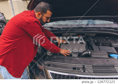 A man repairing his car with the hood open, focused on the engine. Concept of maintenance, auto repair, responsibility, and hands-on work. Everyday moment of problem-solving and care for his vehicle 126772125