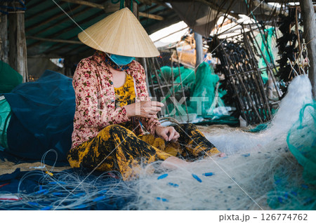 Vietnamese woman in traditional straw hat repairs a fishing net in a workshop on beach in village in Vietnam Vietnamese woman in traditional straw hat repairs a fishing net in a workshop on beach in village in Vietnam 126774762