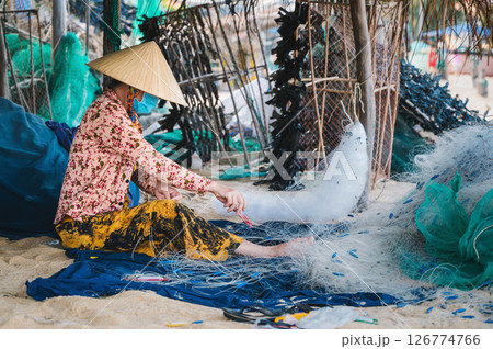 Vietnamese female in straw hat Non La repairs fishing net in a workshop on beach in a fishing village in Vietnam 126774766
