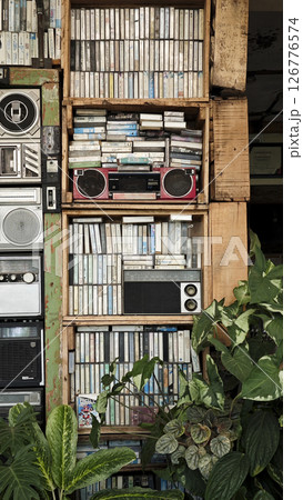 Vintage wooden shelf filled with stacked cassette tapes and retro boomboxes in a rustic setting with wooden crates, green plants, and a nostalgic atmosphere. 126776574