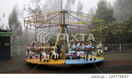 Colorful Carousel with White Horses in a Foggy Outdoor Amusement Park Surrounded by Greenery and Yellow Flowers Colorful Carousel with White Horses in a Foggy Outdoor Amusement Park Surrounded by Greenery and Yellow Flowers 126776647