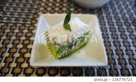 Indonesian Traditional Green Lumpang Cake with Grated Coconut Garnish Served on a White Plate Against a Textured Woven Background 126776721