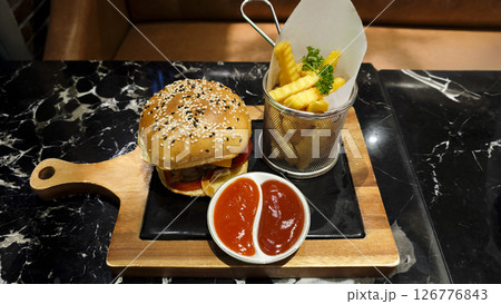 Juicy beef burger with sesame seed bun served on a wooden tray with crispy French fries and dipping sauces on a marble table in a cozy restaurant setting 126776843