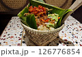 Traditional Nasi Bakul with red chili sambal, fried shallots, and petai served in a woven bamboo basket lined with banana leaves on a colorful terrazzo table in an Indonesian dining setting 126776854
