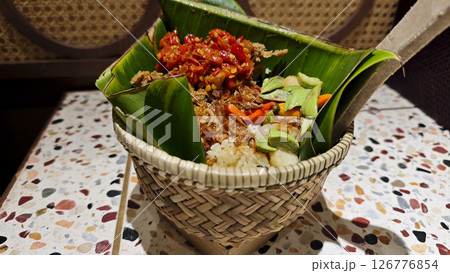 Traditional Nasi Bakul with red chili sambal, fried shallots, and petai served in a woven bamboo basket lined with banana leaves on a colorful terrazzo table in an Indonesian dining setting 126776854