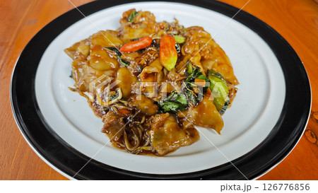 Stir-fried flat rice noodles with rich soy sauce, fresh vegetables, and red chilies served on a white plate with a black rim on a wooden table in a vibrant setting. 126776856