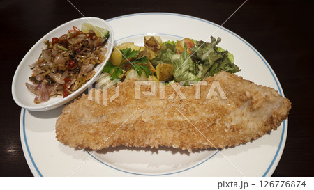 Crispy breaded fish fillet served with sauteed chili onions roasted potatoes fresh green salad and parsley garnish on a white plate creating a vibrant and appetizing dish presentation. 126776874