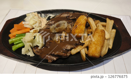 Grilled beef steak served with creamy brown gravy crispy potato wedges french fries and sauteed vegetables on a black sizzling plate creating a delicious hearty meal presentation. 126776875