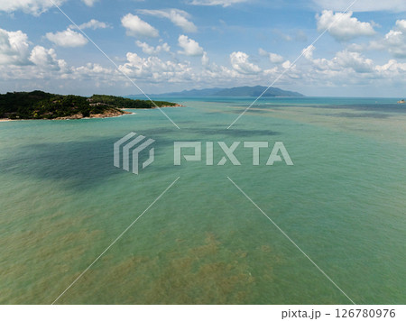 Coastal area with turquoise waters, shadowy patches, and distant mountains under a clear sky with white clouds. Koh Samui, Thailand. Coastal area with turquoise waters, shadowy patches, and distant mountains under a clear sky with white clouds. Koh Samui, Thailand. 126780976