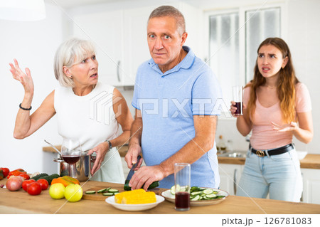 An adult daughter, together with an elderly mother, scolds an elderly father while he is preparing dinner in kitchen An adult daughter, together with an elderly mother, scolds an elderly father while he is preparing dinner in kitchen 126781083