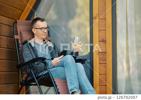 Man looking at smartphone sitting on terrace of a house with a glass of wine 126782087