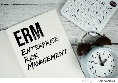 ERM Enterprise Risk Management text on paper notebook page with alarm clock and calculator on wooden background 126782634