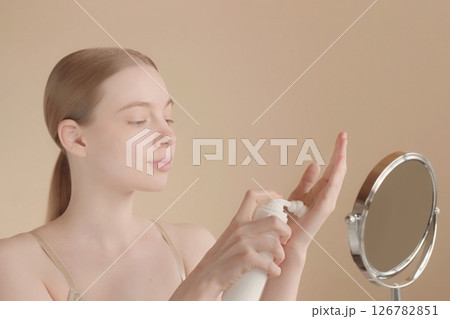 Face. Young woman applying skincare foam on her hand in front of small mirror on isolated light background. Beautiful model girl. Perfect clear skin. Selfcare concept, beauty cosmetics advertisement 126782851