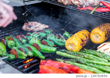 Grilling chicken, corn cobs, paprika, padron, asparagus with temperature control. Meat tongs in human hands. 126783650