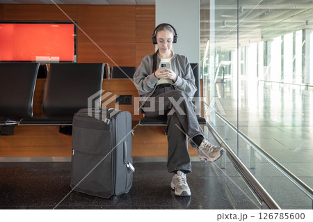 A young woman sits on a bench in a bright airport terminal, wearing headphones and looking at her smartphone with suitcase nearby. Waiting during international travel and digital technology. A young woman sits on a bench in a bright airport terminal, wearing headphones and looking at her smartphone with suitcase nearby. Waiting during international travel and digital technology. 126785600