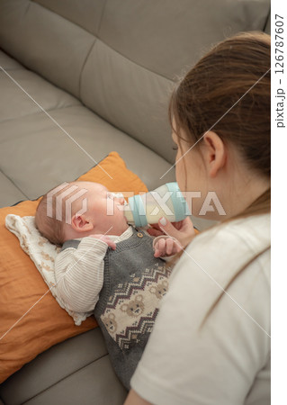 A young mother is bottle-feeding her baby on a cushion on a sofa at home. Concept of infant care, nurturing, early motherhood, feeding routine, and emotional connection in a home setting. A young mother is bottle-feeding her baby on a cushion on a sofa at home. Concept of infant care, nurturing, early motherhood, feeding routine, and emotional connection in a home setting. 126787607