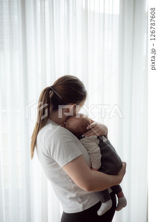 Young mother holding newborn baby in soft natural light standing by a window. Concept of early motherhood, tenderness, bonding, parental love, and infant care in a calm home environment. Young mother holding newborn baby in soft natural light standing by a window. Concept of early motherhood, tenderness, bonding, parental love, and infant care in a calm home environment. 126787608