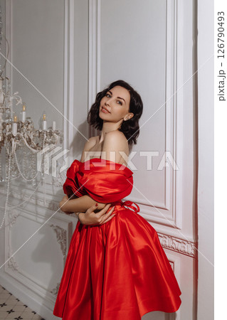 Woman in red off-shoulder dress posing by chandelier in classic white room Woman in red off-shoulder dress posing by chandelier in classic white room 126790493