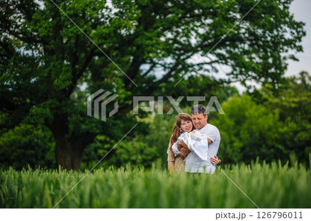Loving parents embracing swaddled newborn near leafy tree, soft sunlight illuminating tender family connection in lush green meadow Loving parents embracing swaddled newborn near leafy tree, soft sunlight illuminating tender family connection in lush green meadow 126796011