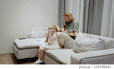 Caring mother gently brushing daughter's hair, sharing tender bonding moment on living room sofa, expressing maternal love and connection Caring mother gently brushing daughter's hair, sharing tender bonding moment on living room sofa, expressing maternal love and connection 126796442