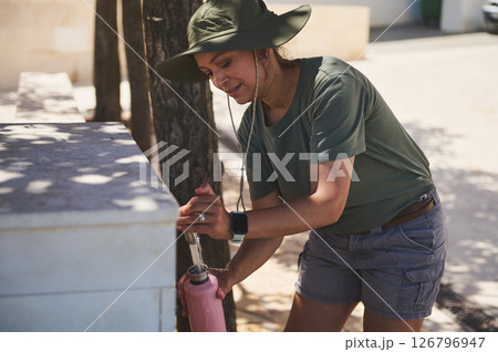 Woman in Hat Filling Water Bottle Outdoors by a Fountain Under Bright Sunlight Woman in Hat Filling Water Bottle Outdoors by a Fountain Under Bright Sunlight 126796947